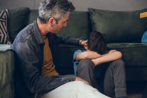 Parent and child sit on the ground. The parents hand on the child's back.