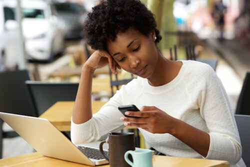A person sitting down by their laptop and using their phone.