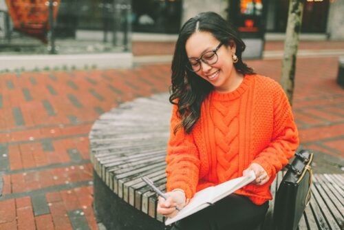 A girl sitting down outside and writing in a notebook.