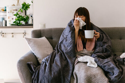 Woman sitting on the couch, blowing her nose with the flu during flu season.