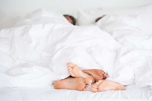 A couple's feet while they lay in bed and talk about the importance of consent.