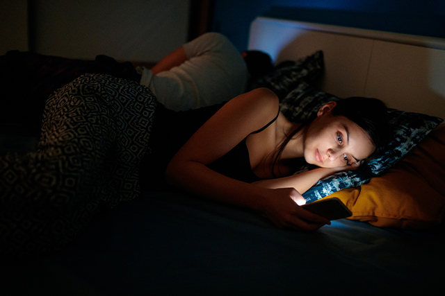 A woman on her phone in bed, representing an activity she should avoid before bed.