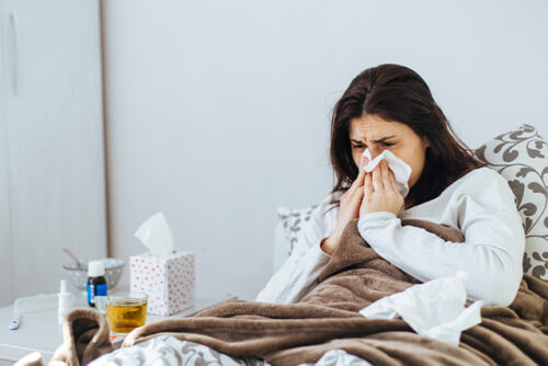 A woman blowing her nose on a tissue wondering difference between cold and flu and which illness she has.