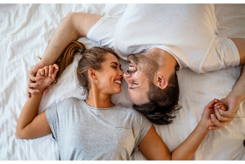 A couple smiling while laying in bed and holding hands.
