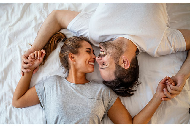 A couple smiling while laying in bed and holding hands.
