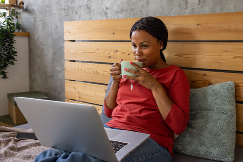 woman in bed sipping coffee and looking at laptop