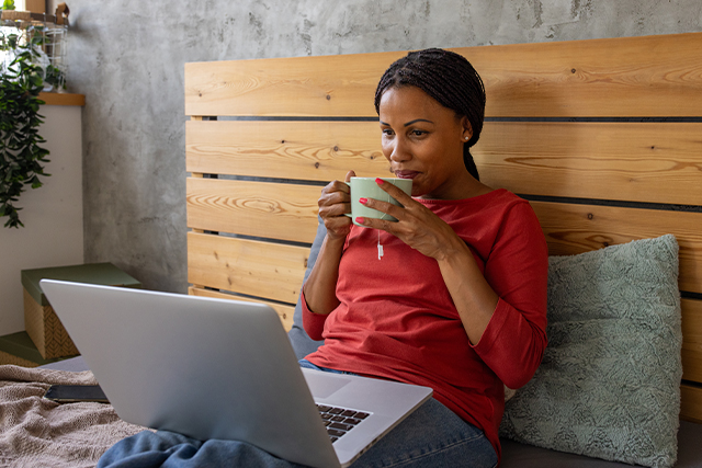 woman in bed sipping coffee and looking at laptop