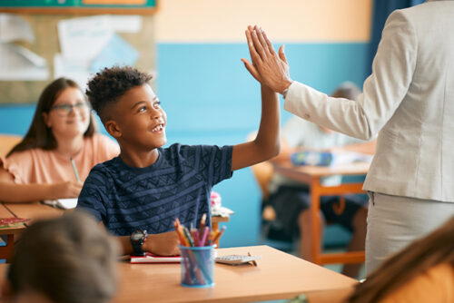 Nine year old sitting at a desk high fives a teacher passing by.