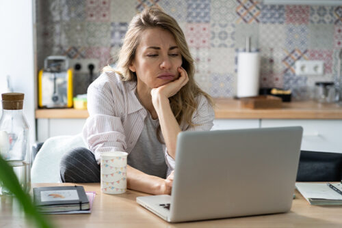 Woman sits at table looking at computer with her chin in her hand.