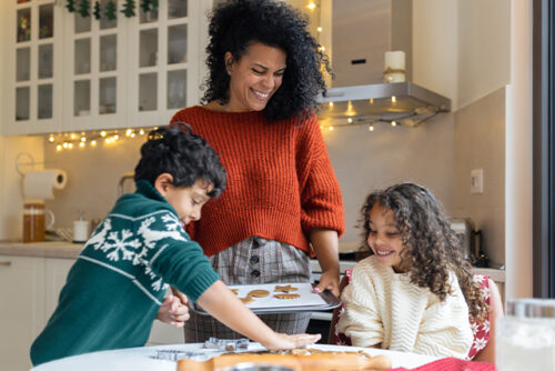 Two kids sit at the kitchen table, making cookies. A parent stands next to them smiling with a cookie baking sheet.