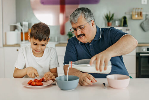 Father and son sit at counter eating breakfast. Father pours milk into his son's bowl.
