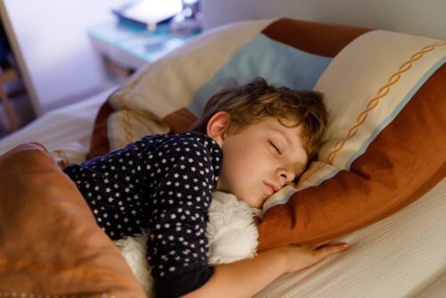 Child sleeps in bed facing the camera and cuddling a stuffed animal