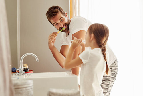 Father and daughter stand on the bathroom looking at each other as they brush their teeth
