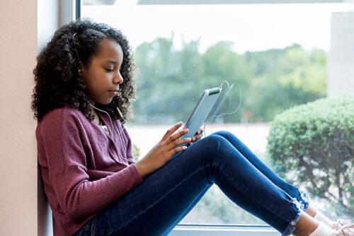 Kid sits in the window, reading from an ipad with headphones in.