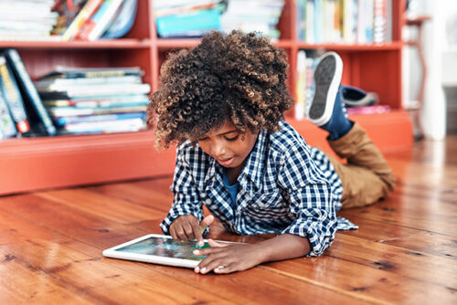 Child lays on ground on their stomach reading from an ipad.
