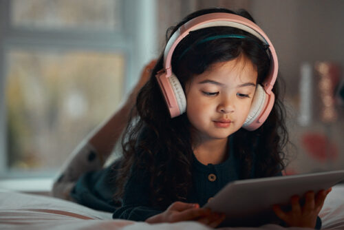 Child with headphones on lays in bed looking at ipad.
