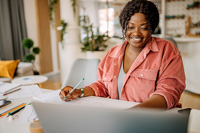 Woman holding pen, looks at computer screen smiling.