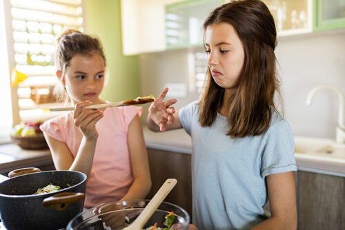 Two children stand in the kitchen looking disgusted at a spoon of food.