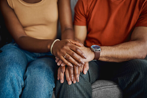 Two people sit on a couch, their faces out of view, holding hands.
