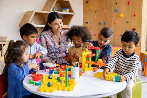 Teacher sits with young students around a table playing