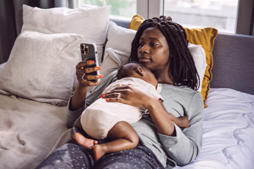 Woman in bed holding baby while looking at mobile phone