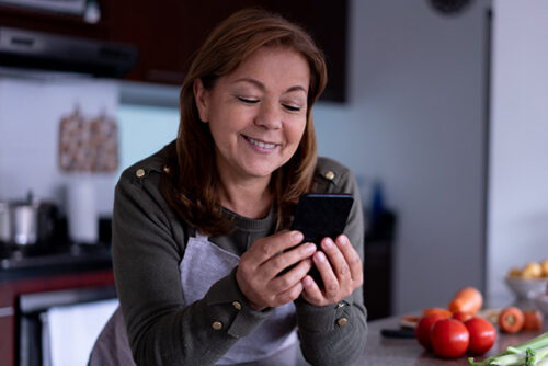 Woman stands in kitchen, leaning over the counter and smiling at her phone.