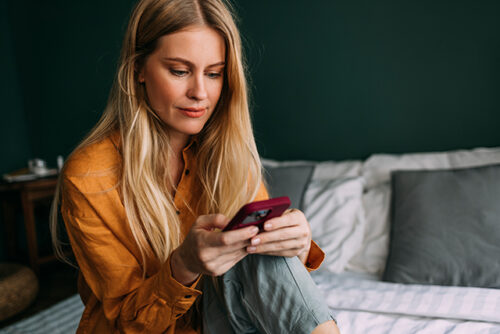 Woman sits on bed, looking at phone contemplatively.
