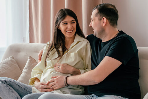 Pregnant woman and man look at each other smiling with their hands on her baby bump.