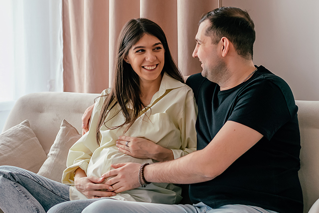 Pregnant woman and man look at each other smiling with their hands on her baby bump.