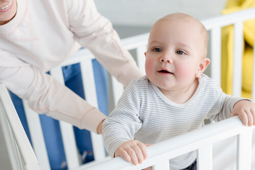 baby stands up in crib with support from an adult