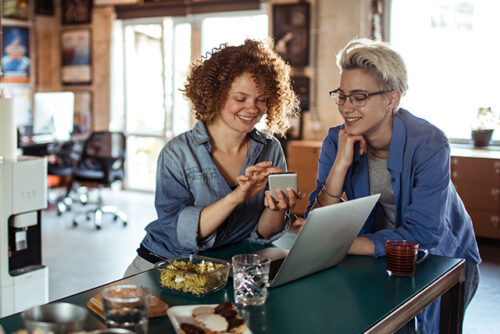 Two women sit at the kitchen table looking at a phone and smiling