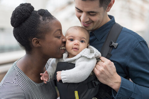 man holds baby in carrier, woman kisses baby