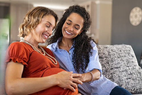 two women cuddle on the couch, one holds her bump