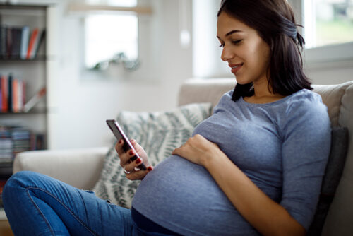 A woman looks down at her phone while holding her baby bump