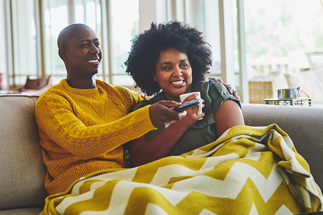 two people sit on a couch under a blanket, the man points the remote control at a TV out of sight.