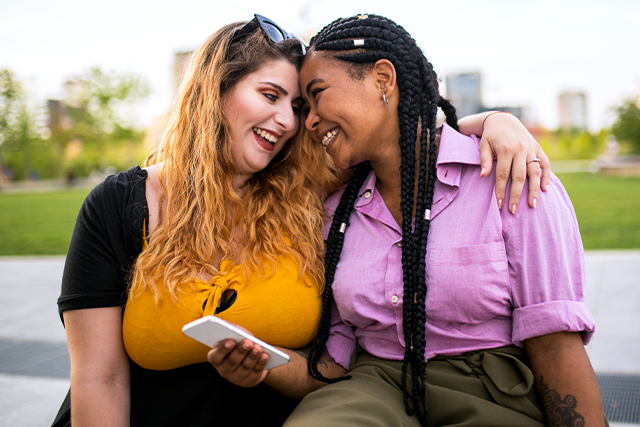 Two women laugh while sitting on a park bench with their arms around each other