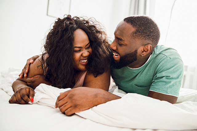Two people lie on their stomachs on a bed smiling at each other