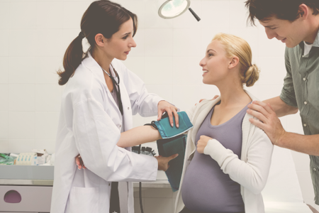 pregnant woman with partner in doctor's office with doctor strapping blood pressure cuff around her arm