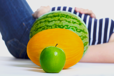a pregnant woman laying next to a series of fruits the size of her stomach