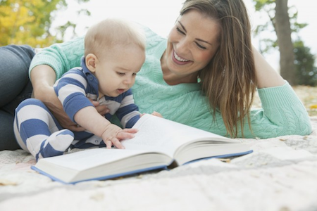 woman lying with baby outside on a picnic blanket reading a book