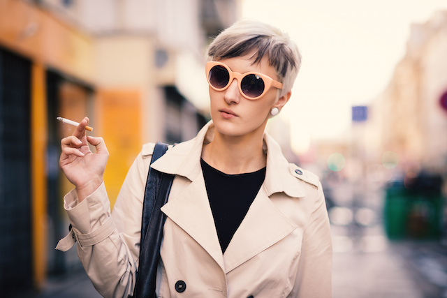 A person with sunglasses on walking and holding a cigarette.
